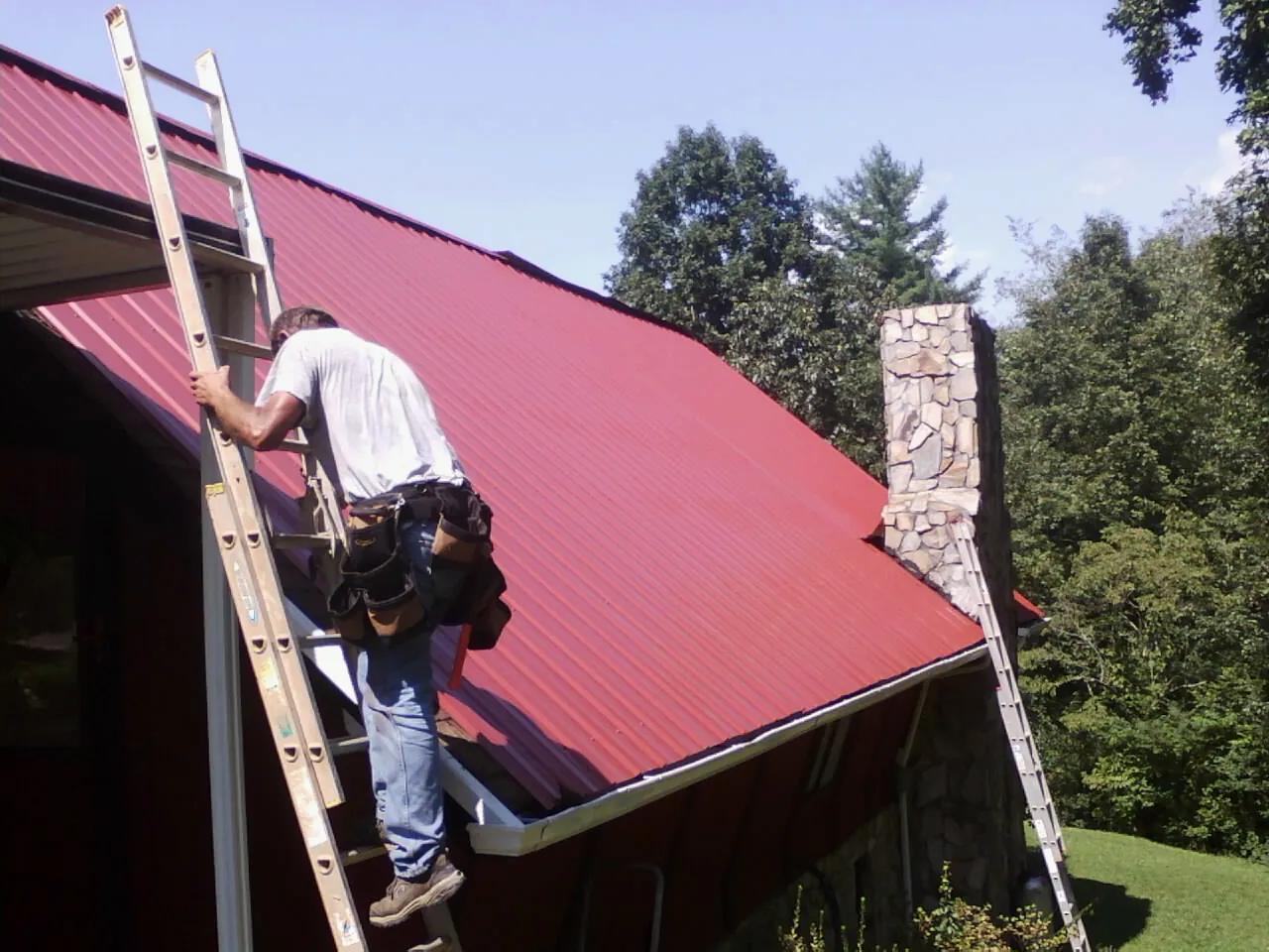 Roofer on a ladder beside a red metal roof and stone chimney — Soffit & Fascia Repair by Roofing Brothers in Ansted